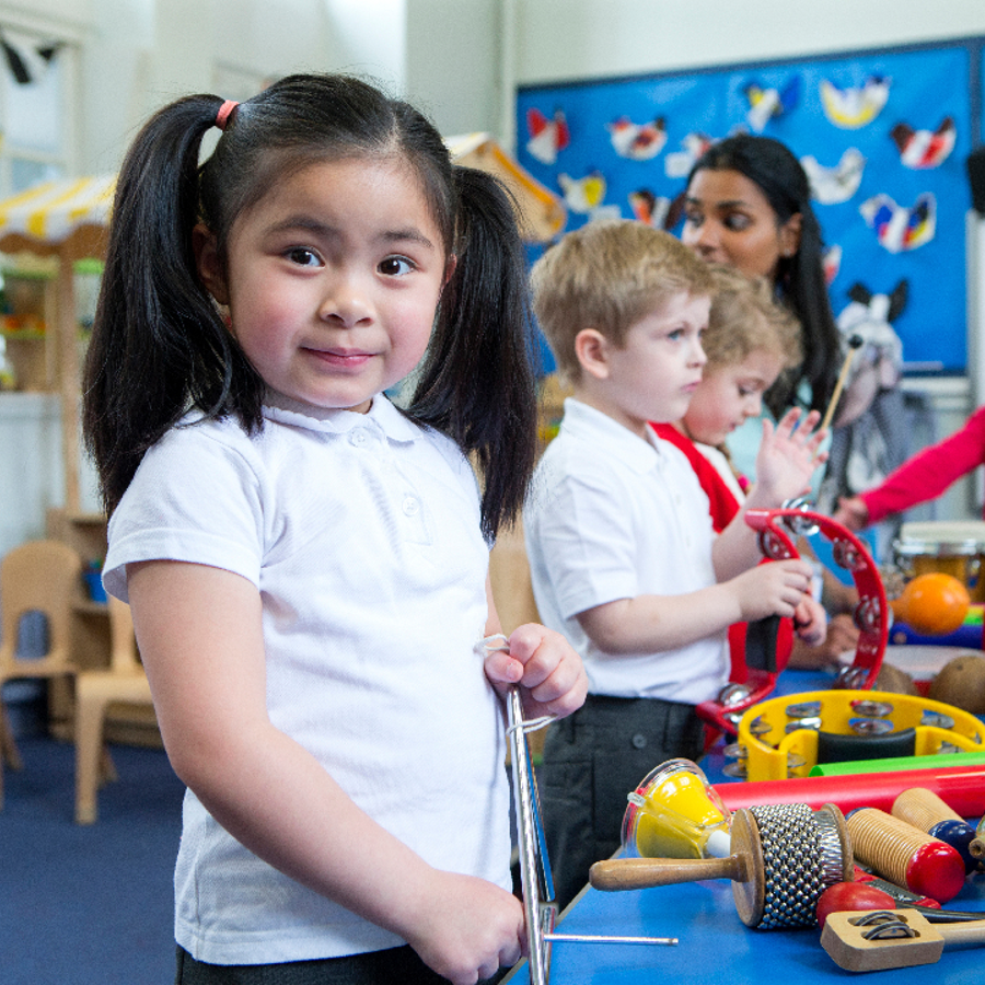 Primary school children in school uniform standing around a table playing with different musical instruments. A young girl in the foreground is staring directly into the camera.