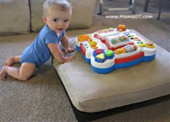 Baby leaning on sofa cushion on the floor. A colourful toy is on top of the sofa cushion