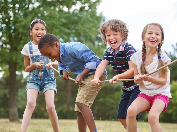 Four children pulling a rope in a game of tug of war whilst laughing and smiling outside in a park.