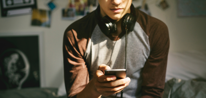 Teenage boy sitting on bed with headphones round their neck, looking on their phone.