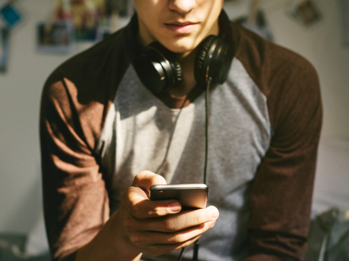 Teenage boy sitting on bed with headphones round their neck, looking on their phone.