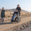 A child being pushed along in a wheelchair by an adult along a sandy path with another young girl walking a dog beside them