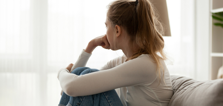 Teenage girl sitting on the sofa, hugging her knees to her chest with one arm and resting her other fist on her face. She is staring out the window away from the camera.