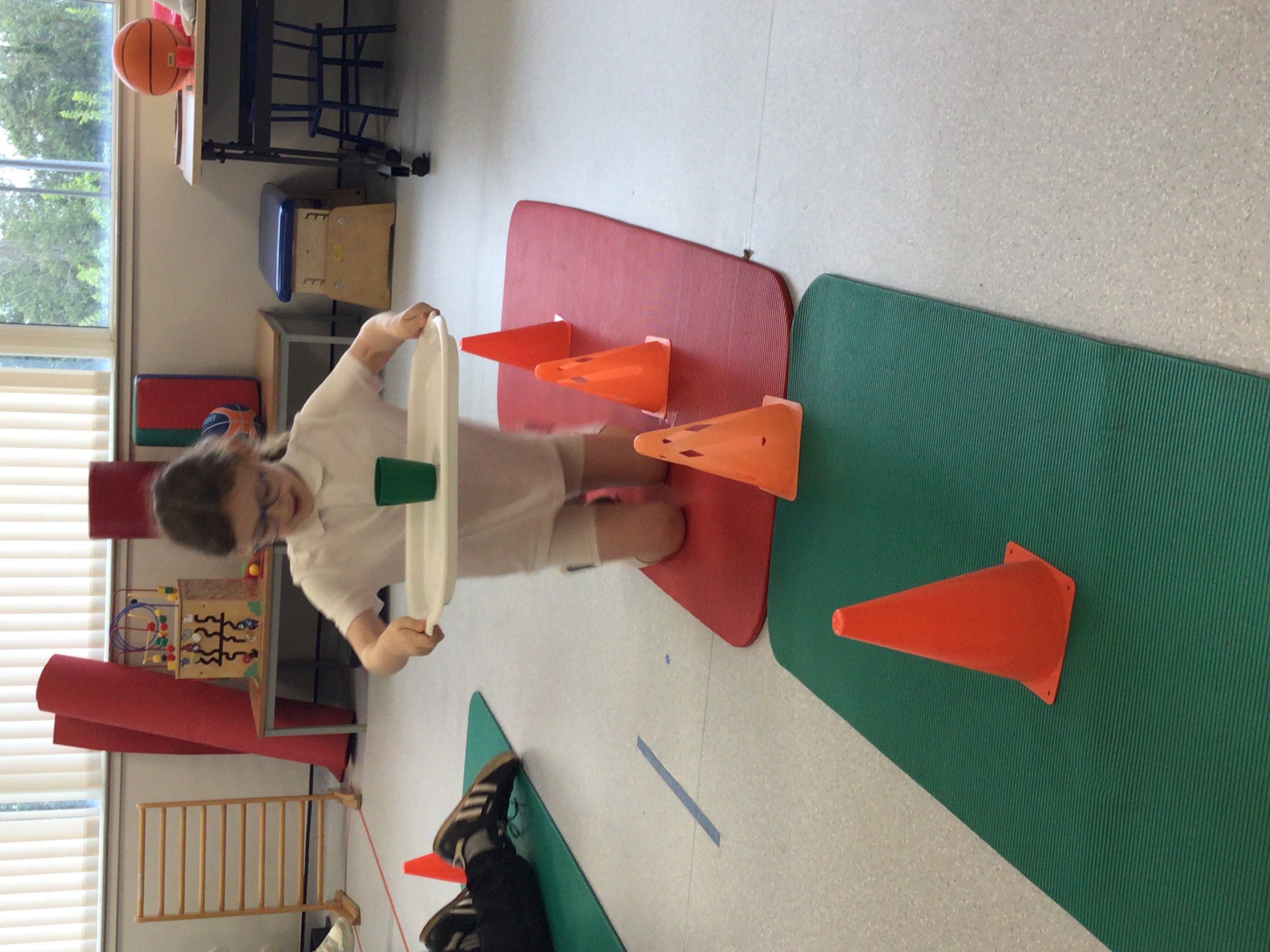 Child carrying a tray with a glass ull of water walks on her knees around some cones on the floor