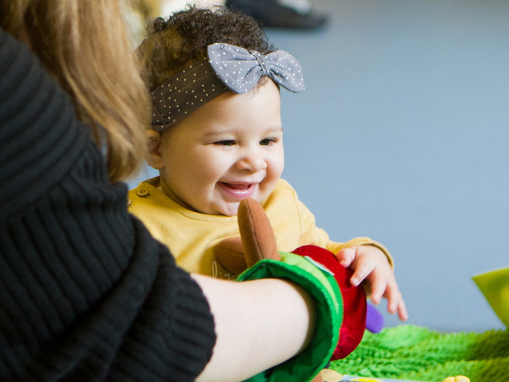 Young baby sitting on the floor smiling as her mum is reaching round her with a hand puppet.