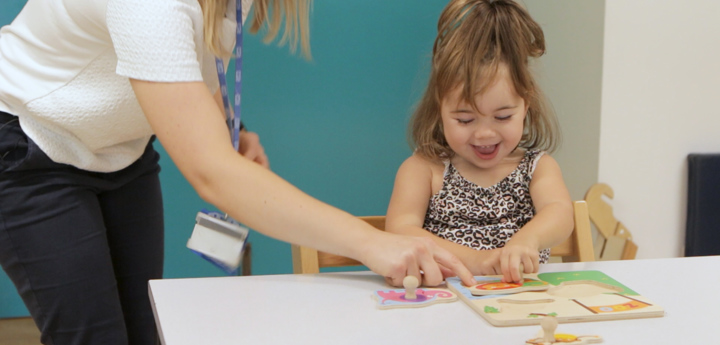 Young girl playing with a puzzle and smiling whilst an adult is pointing at a puzzle piece