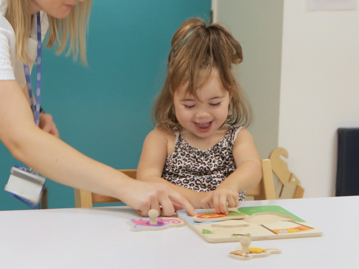 Young girl playing with a puzzle and smiling whilst an adult is pointing at a puzzle piece