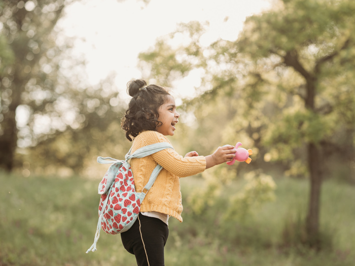 Little girl with backpack standing in a forest holding a pink toy and smiling