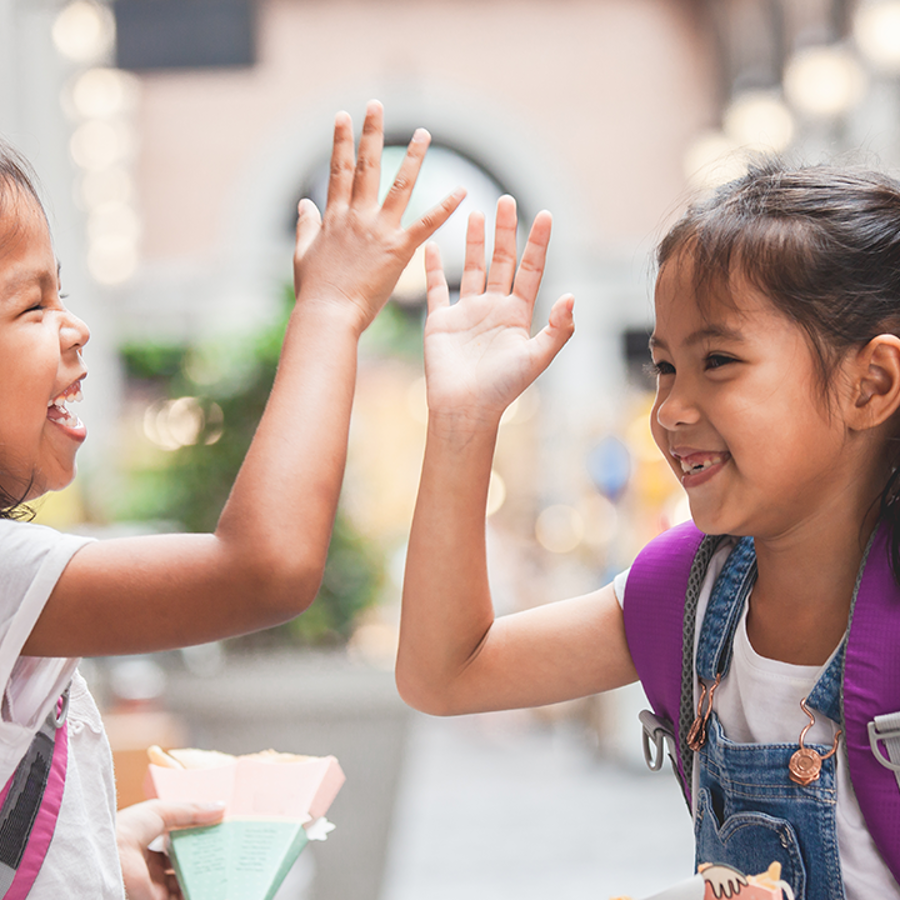 2 young girls wearing backpacks high-fiving whilst smiling and laughing