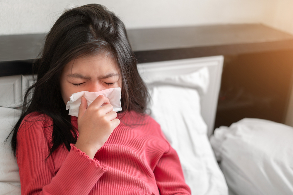 Girl sitting up in bed sneezing into a tissue.