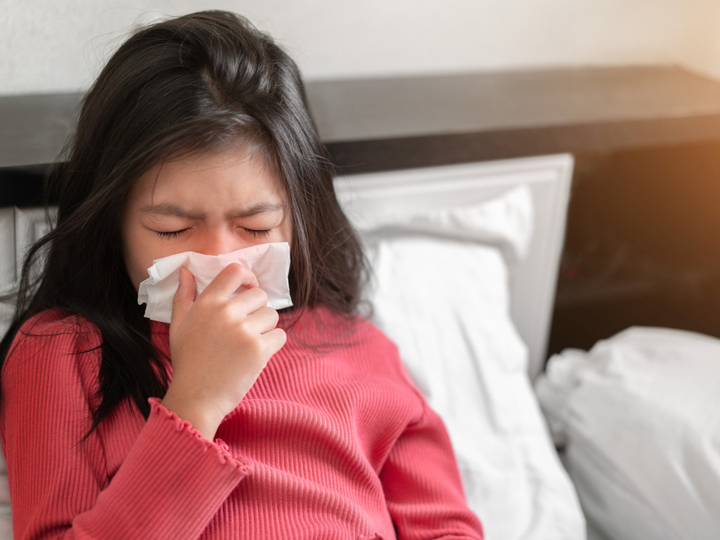 Girl sitting up in bed sneezing into a tissue.