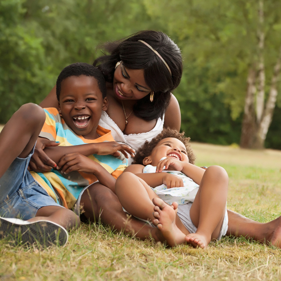 Mum hugging two children and laughing whilst sitting on the grass at a park.