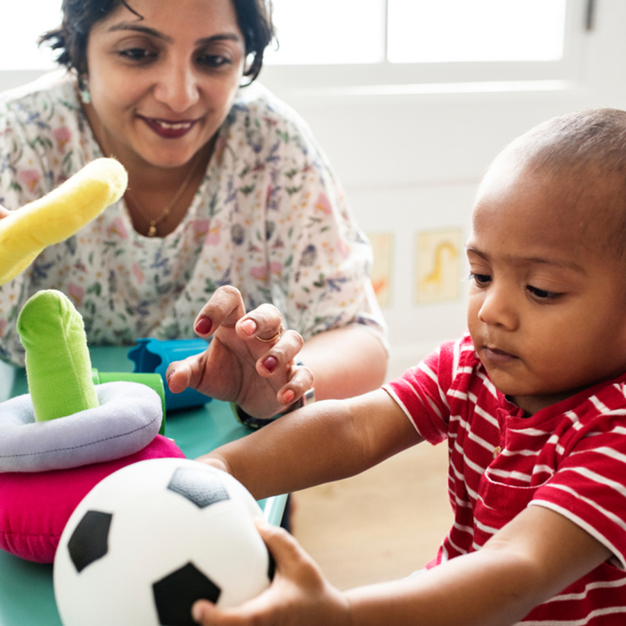Toddler sitting at a child's table holding a football. A smiling adult is also sitting at the table and playing with the other soft toys on the table. 