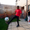 Teenage Boy Playing Kicking Ball In Back Garden on a stone patio with their male friend behind filming on their phone.