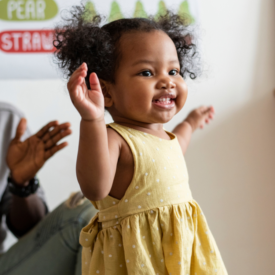 Girl toddler dancing with her hands up in the air, with her dad smiling and clapping behind her.