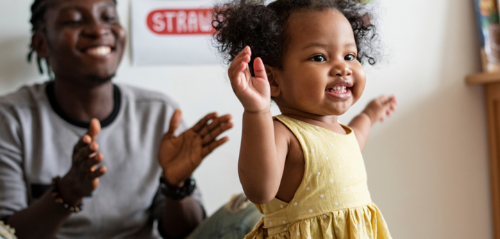 Girl toddler dancing with her hands up in the air, with her dad smiling and clapping behind her.