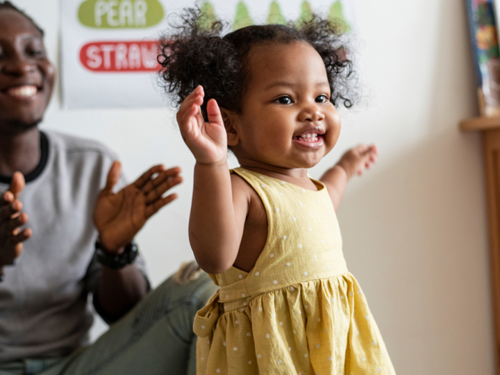 Girl toddler dancing with her hands up in the air, with her dad smiling and clapping behind her.