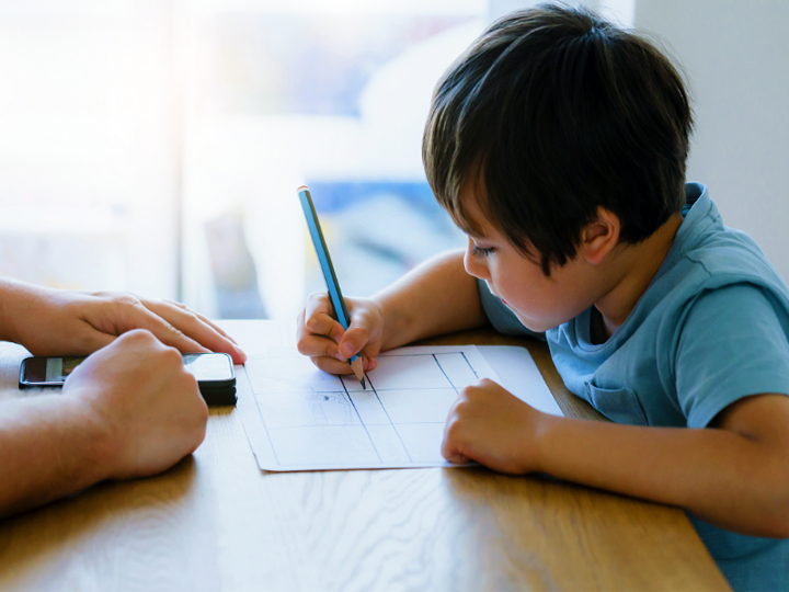 Young boy sitting at a table writing on a piece of paper with an adult sitting opposite of them.