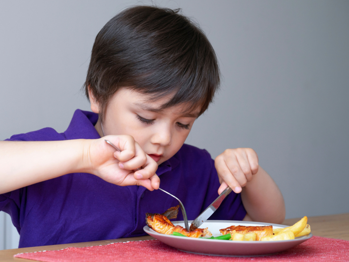 Boy sitting at table and using knife and fork to eat their food off their plate. 