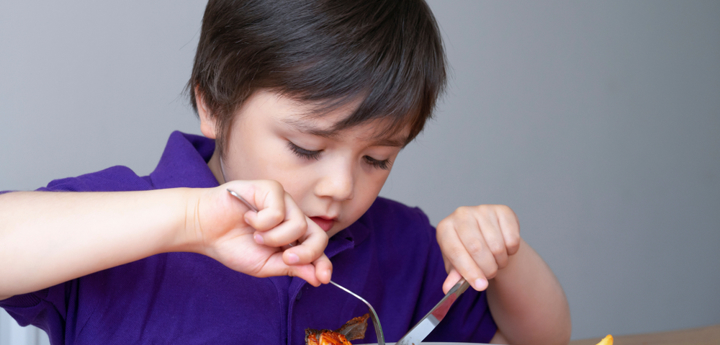 Boy sitting at table and using knife and fork to eat their food off their plate. 