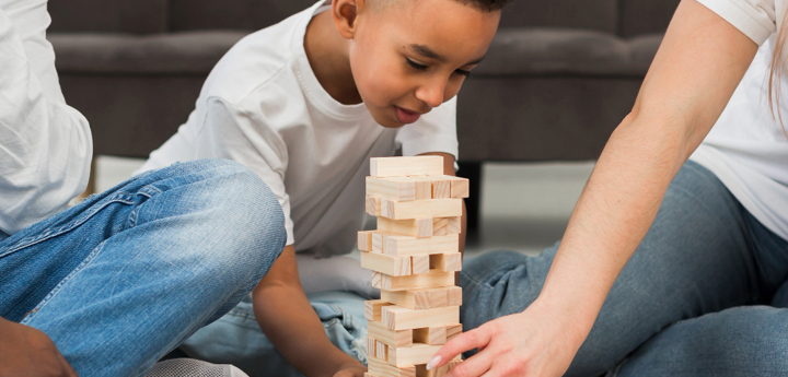 Boy playing Jenga with 2 adults either side of him. All sitting on the floor.