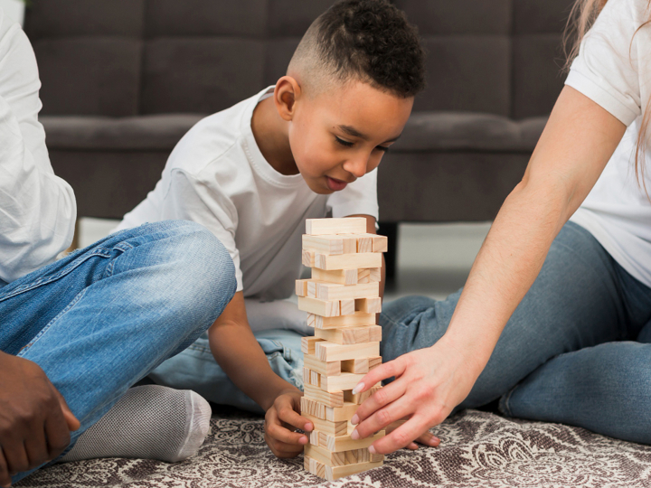 Boy playing Jenga with 2 adults either side of him. All sitting on the floor.