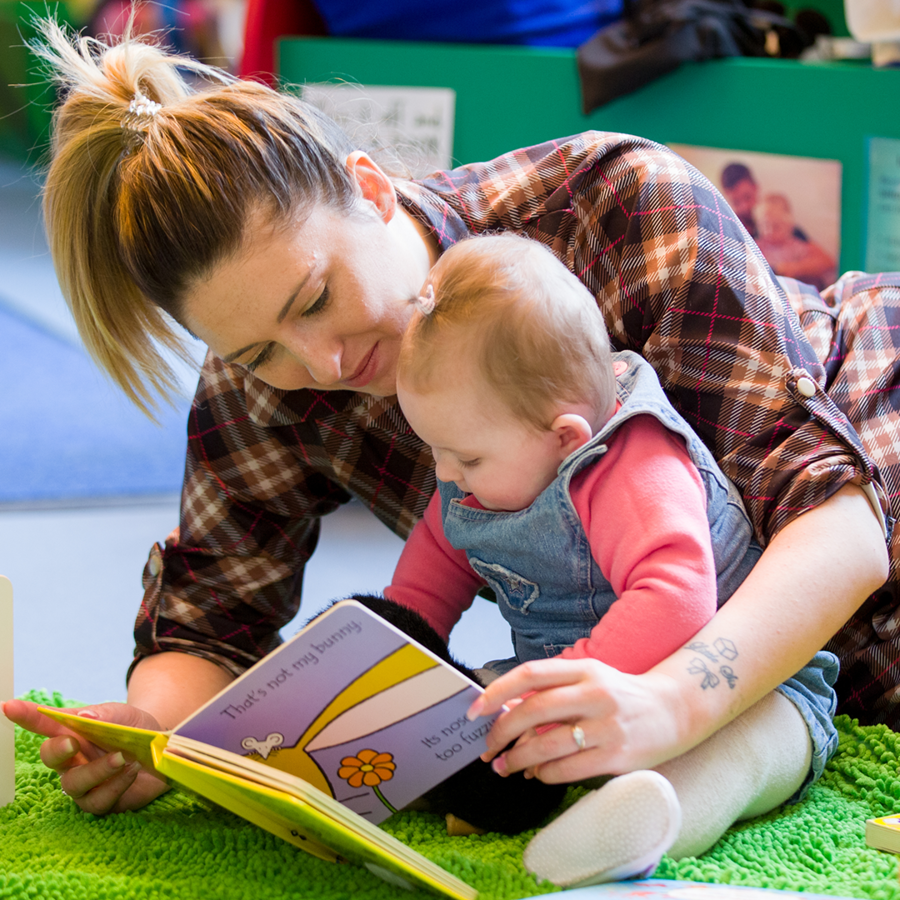 Adult leaning over baby girl who is sitting up and reading a book to her.