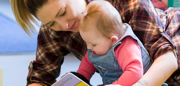 Adult leaning over baby girl who is sitting up and reading a book to her.
