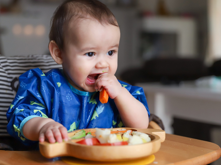 Baby boy sitting in highchair holding a carrot stick to his mouth with a plate of mixed vegetables in front of him. 