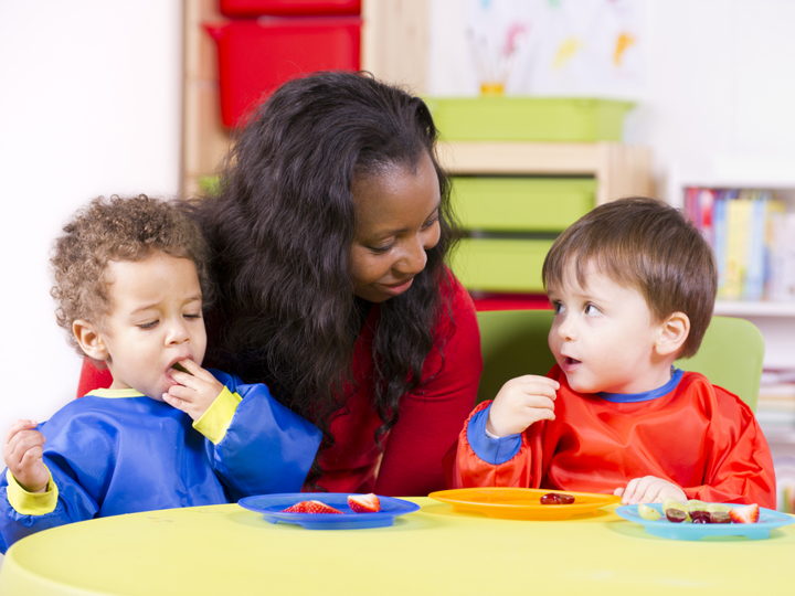 Two toddlers eating fruit sitting at a child's table in a nursery setting. A carer is sitting behind them.