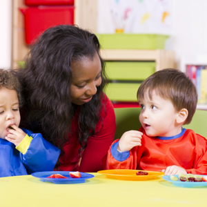Two toddlers eating fruit sitting at a child's table in a nursery setting. A carer is sitting behind them. 