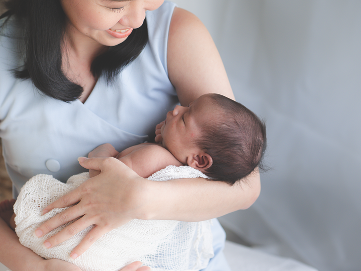 A woman sitting down and smiling whilst looking down at the baby she is holding, which is wrapped in a blanket, in her arms