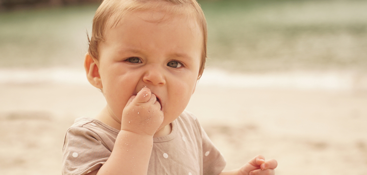 Young baby sitting on the beach, bringing their right hand up to their mouth and eating sand