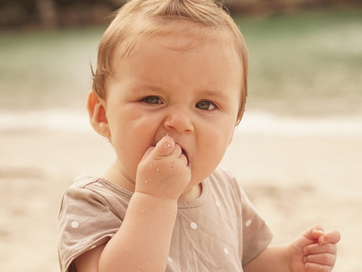 Young baby sitting on the beach, bringing their right hand up to their mouth and eating sand