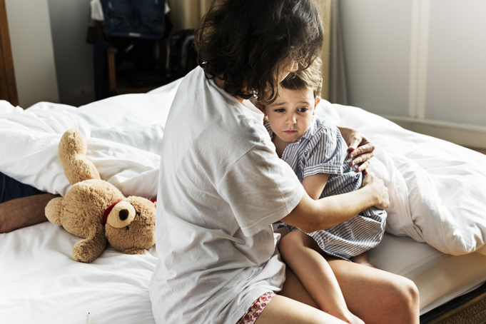 Adult comforting a child looking concerned. On a bed with a teddy bear in the background