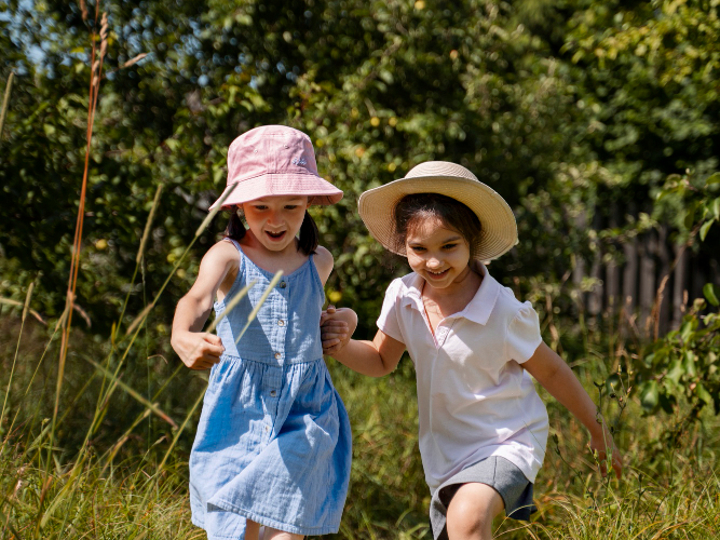 Two young girls running through grass in the sun wearing sun hats