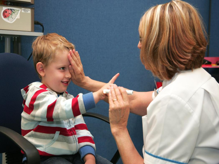 Young boy sitting in a chair with a clinician covering one of his eyes with the palm of her hand and holding a light in the other hand. 