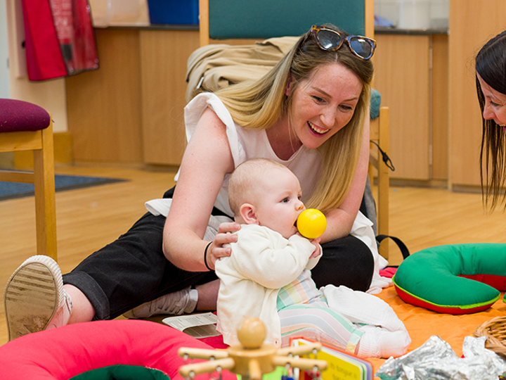 Two adults sitting on the floor interacting with a baby who is also sitting on the floor. They are surrounded by toys.