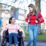 Two young girls talking. One girl is sitting in a wheelchair whilst the other girl is standing next to her with a backpack on.