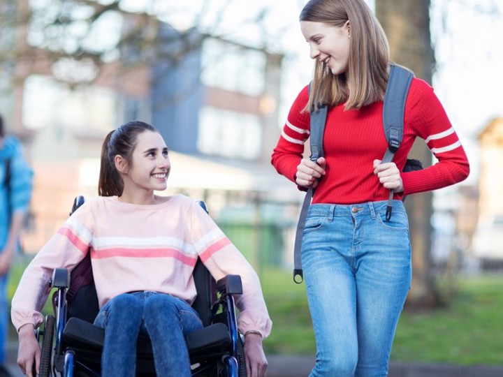 Two young girls talking. One girl is sitting in a wheelchair whilst the other girl is standing next to her with a backpack on.
