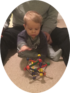 Baby sat in between legs of an adult reaching for a colourful toy