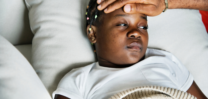 Child lying on sofa under a blanket looking unwell. An adults hand is placed on their forehead, feeling their temperature. 