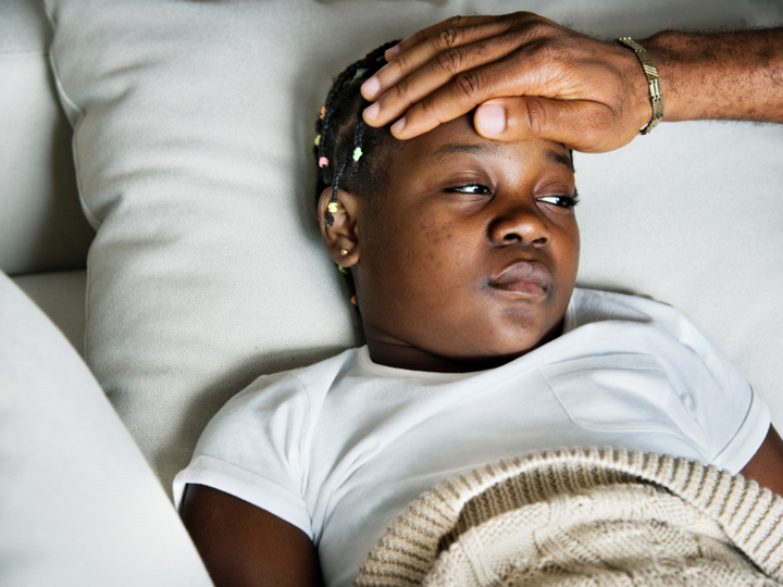 Child lying on sofa under a blanket looking unwell. An adults hand is placed on their forehead, feeling their temperature. 