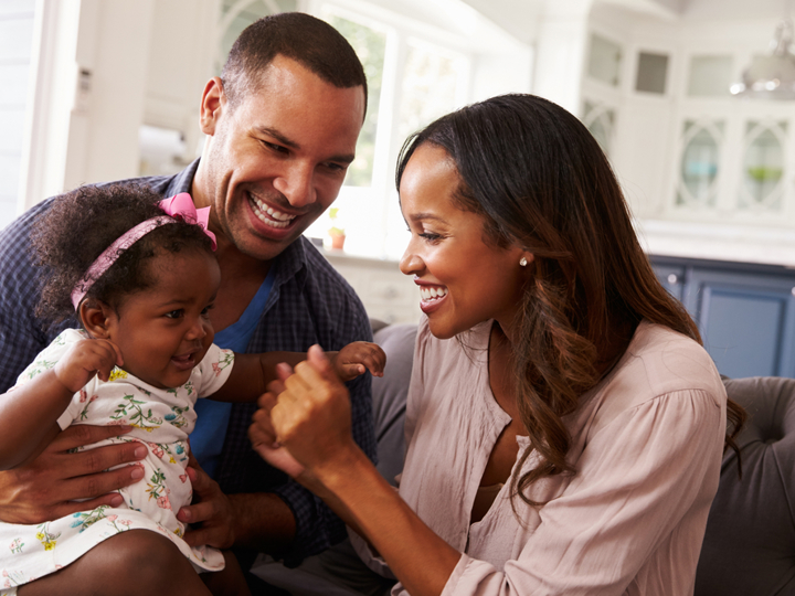 Mum and dad sitting on sofa laughing and smiling with a happy baby girl sitting on the dad's knee