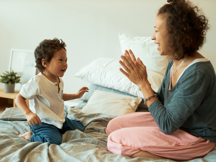 Mum and young son kneeling on the bed together. Both are smiling and look happy whilst mum is clapping her hands