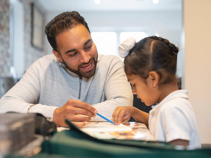 Adult man and child sitting at a table. The man is pointing to a page of a book in front of them with a pencil.