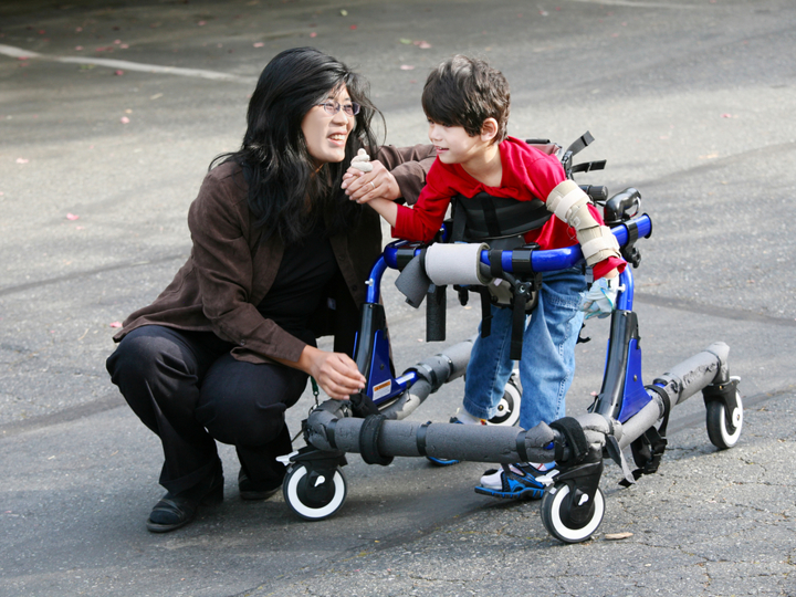 Adult woman crouching next to young boy with walking support. Woman is holding the boys hand.
