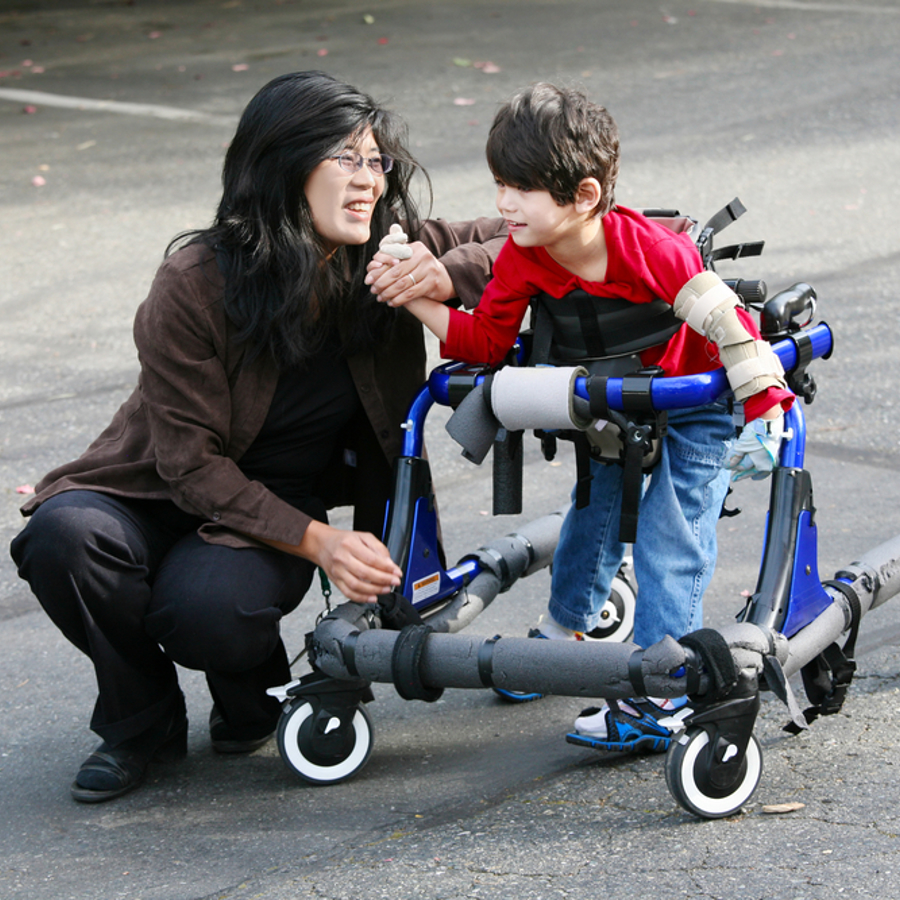Adult woman crouching next to young boy with walking support. Woman is holding the boys hand.