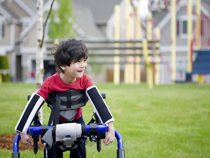 Young boy outside smiling being supported to walk by a walking aid.
