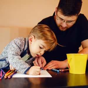 A child drawing with crayons and pencils on a coffee table whilst sitting on a sofa. Next to the child is their dad who is also drawing on the paper